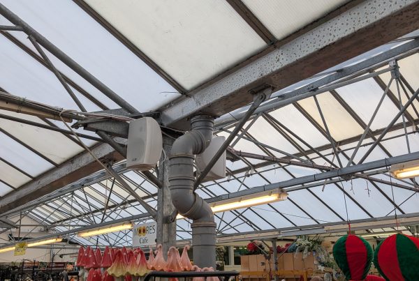 Interior of a garden center or greenhouse with a translucent roof, metal beams, and shelves displaying red mushroom garden ornaments, while a few shoppers browse in the background.
