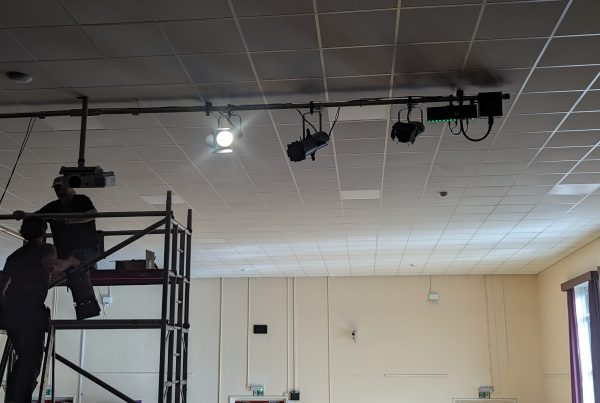 Two people stand on a scaffold adjusting stage lighting equipment mounted to a ceiling rig in a large room, with a projector and multiple lights visible overhead.