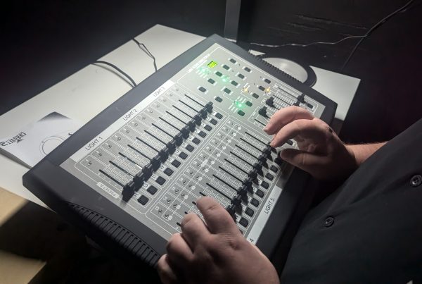 A person adjusts sliders on a professional lighting control console, with multiple labeled channels and illuminated buttons, set on a desk in a dimly lit room.