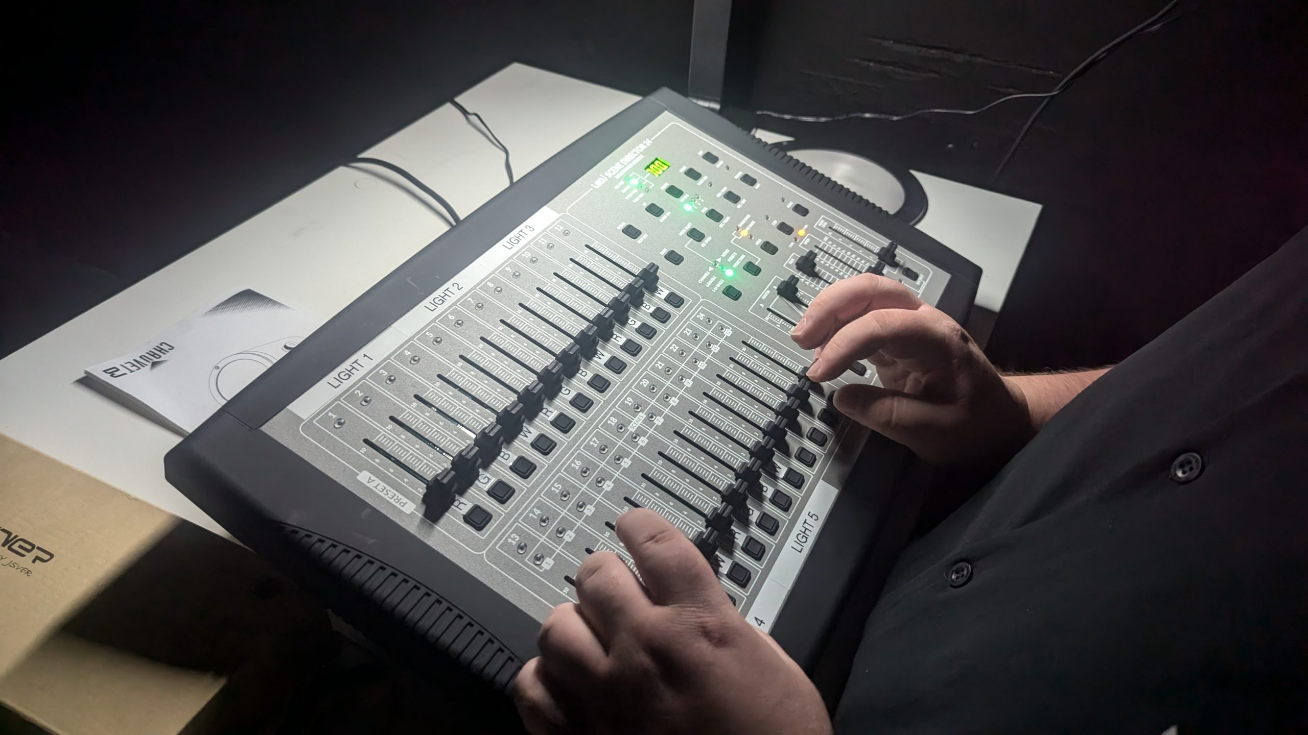 A person adjusts sliders on a professional lighting control console, with multiple labeled channels and illuminated buttons, set on a desk in a dimly lit room.