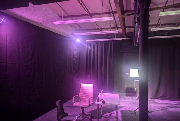 A dim studio space with black curtains and purple lighting, featuring a small table with chairs, a standing floor lamp, and overhead fluorescent fixtures, creating a staged or interview-like setup.