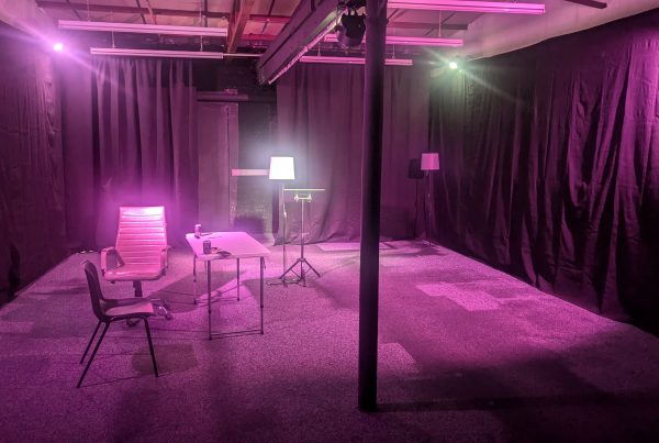 A studio setup with black draped walls and purple-pink lighting, featuring a small table with two chairs, a bright floor lamp on a stand, and a central support column under exposed ceiling fixtures.